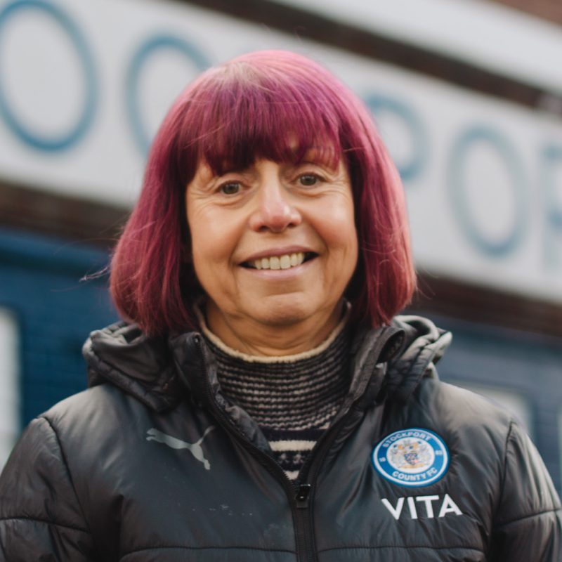A photo of Cath Shanley from Stockport County Supporters Cooperative stood outside the Stockport County sign at Edgeley Park, Stockport.