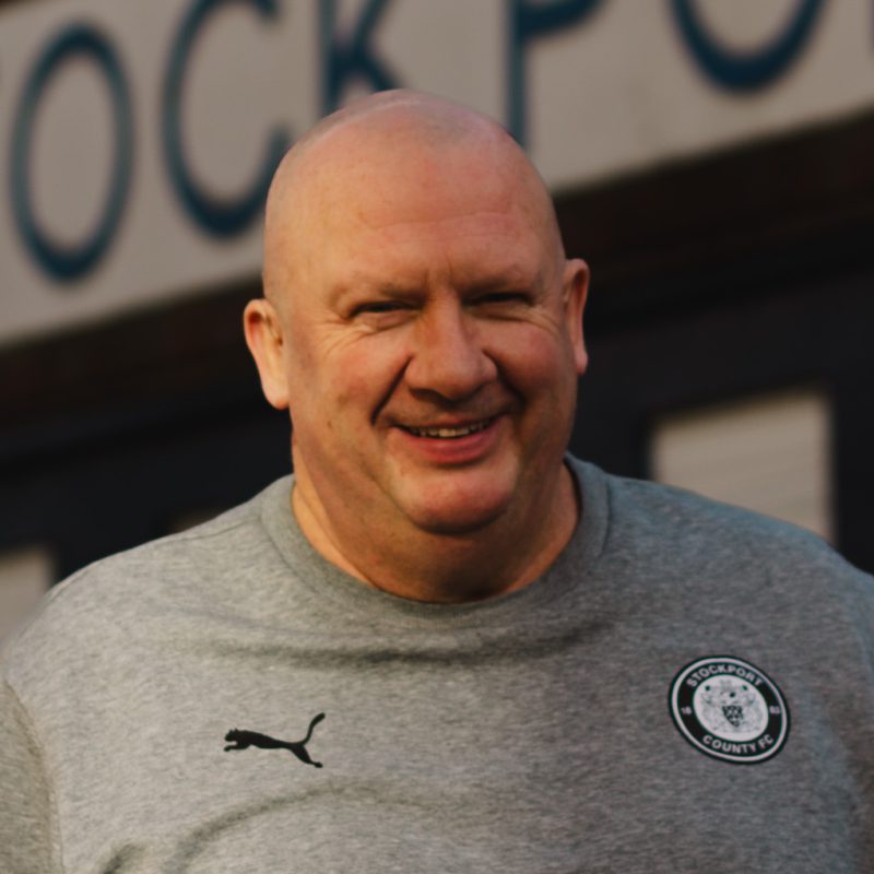 A photo of Dave Marchbank from Stockport County Supporters Cooperative stood outside the Stockport County sign at Edgeley Park, Stockport.