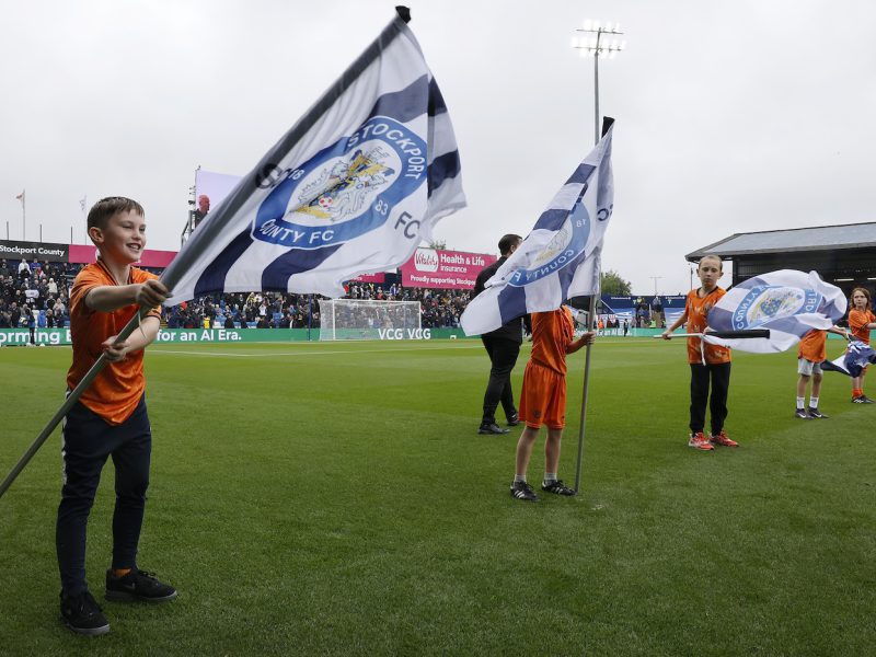 A photograph of the match officials entering the field of play with Children from the Stockport County Match Day Flag Bearer Programme on either side.