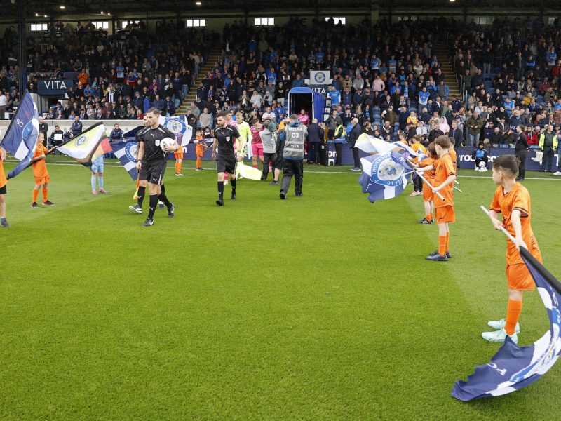 A photograph of the match officials entering the field of play with Children from the Stockport County Match Day Flag Bearer Programme on either side.