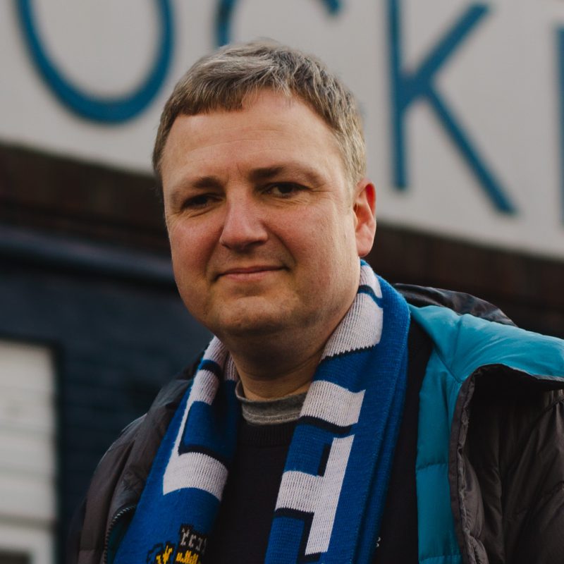 A photo of John Giles from Stockport County Supporters Cooperative stood outside the Stockport County sign at Edgeley Park, Stockport.