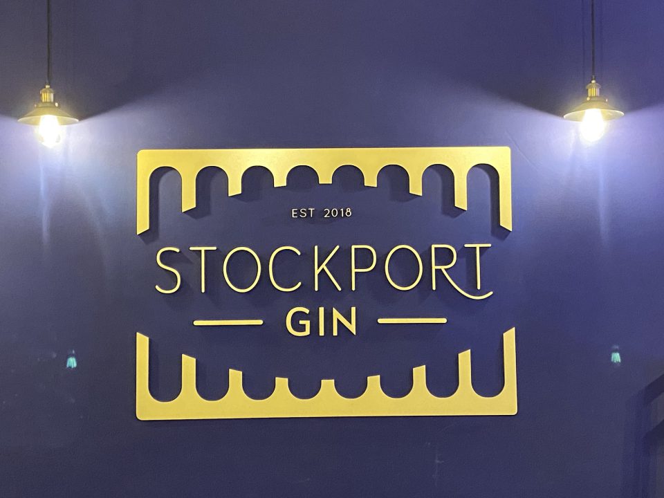 A photograph of the Stockport Gin logo. The logo is yellow on a blue background.